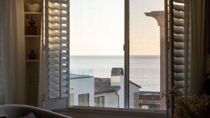 Une pièce donnant sur la mer grâce à des volets ouverts, avec une chaise blanche et une étagère ornée d’objets décoratifs.