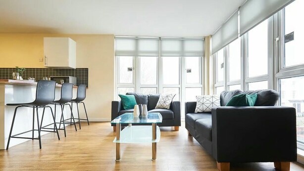 Ett modernt vardagsrum med ett soffbord i glas, två soffor med kuddar och en matplats med stolar. Stora fönster släpper in naturligt ljus och ger utsikt över omgivningen.
