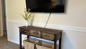 Un téléviseur à écran plat fixé au mur, au-dessus d’une console en bois dotée de tiroirs et de paniers en osier.