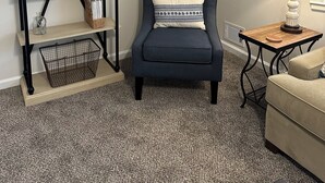 Un salon comprenant un fauteuil bleu foncé, un canapé beige, une table d’appoint en bois avec une lampe, une bibliothèque contenant divers objets et un élément décoratif mural.