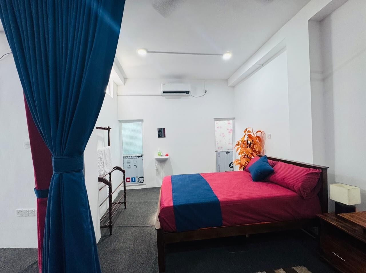 A bedroom with a bed, a desk, and a chair. There is a window with curtains, an air conditioning unit, and a small table with a lamp.