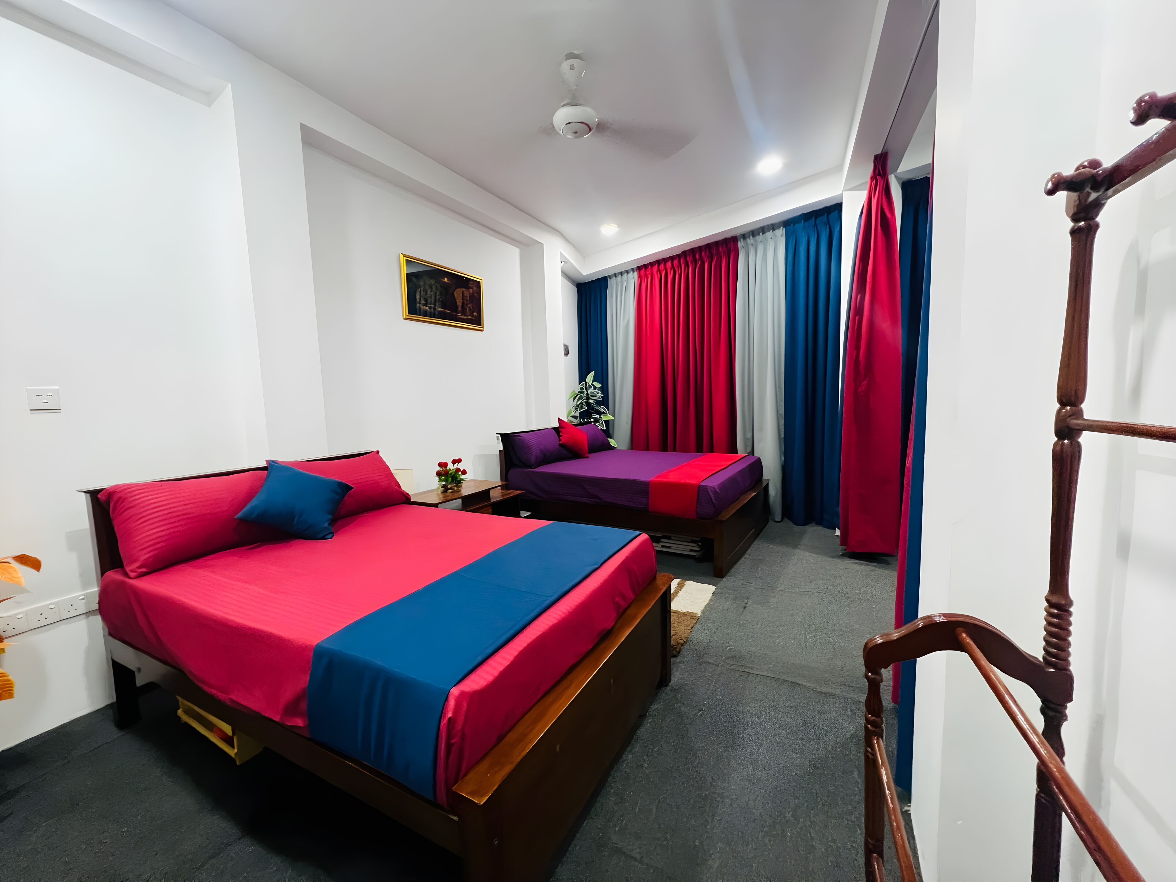 Quarto de hotel com duas camas, ventilador de teto e janela com cortinas vermelhas e azuis.