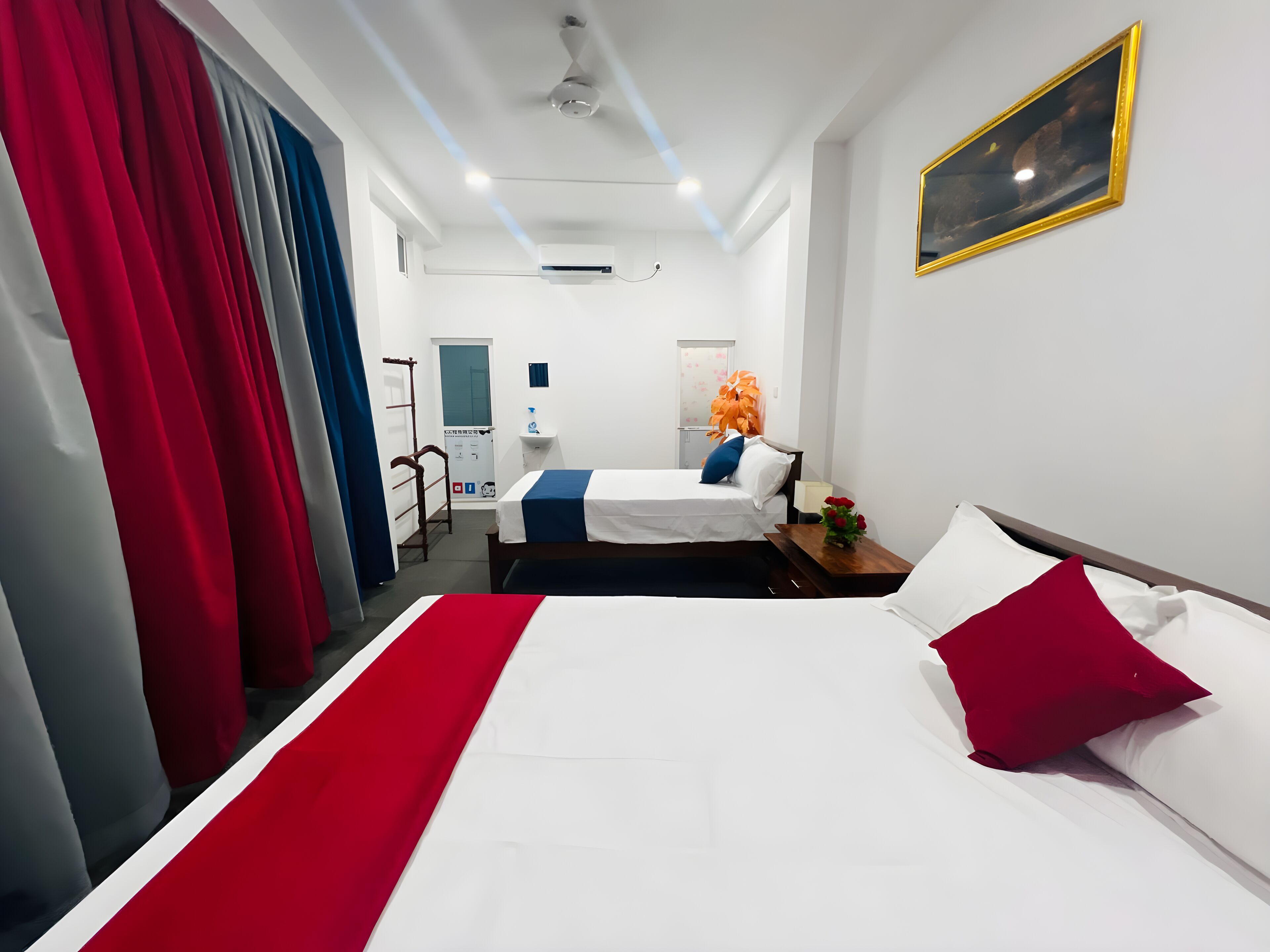 Habitación de hotel con dos camas, una colcha roja y blanca, almohada roja, mesita de noche con un florero y un ventilador de techo, además de aire acondicionado instalado en la pared.