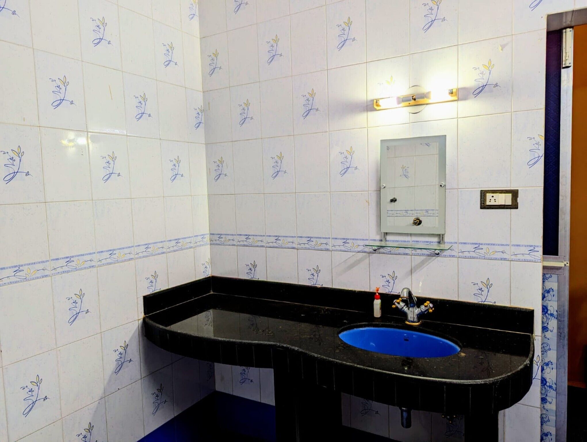 Un bagno con piastrelle bianche e motivi floreali blu, un lavabo nero con una bacinella blu e uno scaffale in vetro sopra il lavabo.