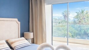 Una habitación de hotel con una cama grande, vista a palmeras a través de la ventana y una decoración de toalla con forma de corazón sobre la cama.