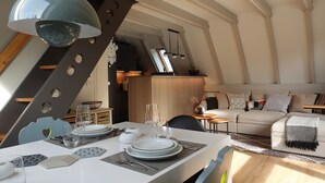 A dining area with a white table set for four, a modern pendant light, and a view of the living room with a sofa and a TV. - Das-Bergheim.de A stylish vacation home in Chiemgau! (Siegsdorf)
