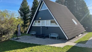 A modern, triangular-shaped building with a sloped roof, a small porch with two benches, and a clear view of the surrounding trees. - Das-Bergheim.de A stylish vacation home in Chiemgau! (Siegsdorf)
