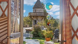 Una puerta abierta que conduce a una torre de piedra con balcón, rodeada de follaje y un sendero de piedra.