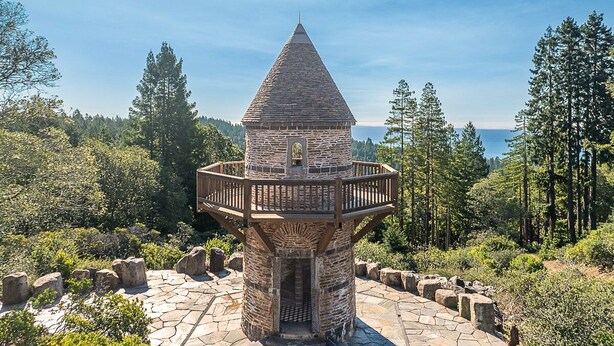 Una torre de observación de piedra con balcón de madera, rodeada de árboles y un cielo despejado.
