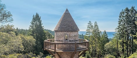 Una torre de observación de piedra con balcón de madera, rodeada de árboles y un cielo despejado.