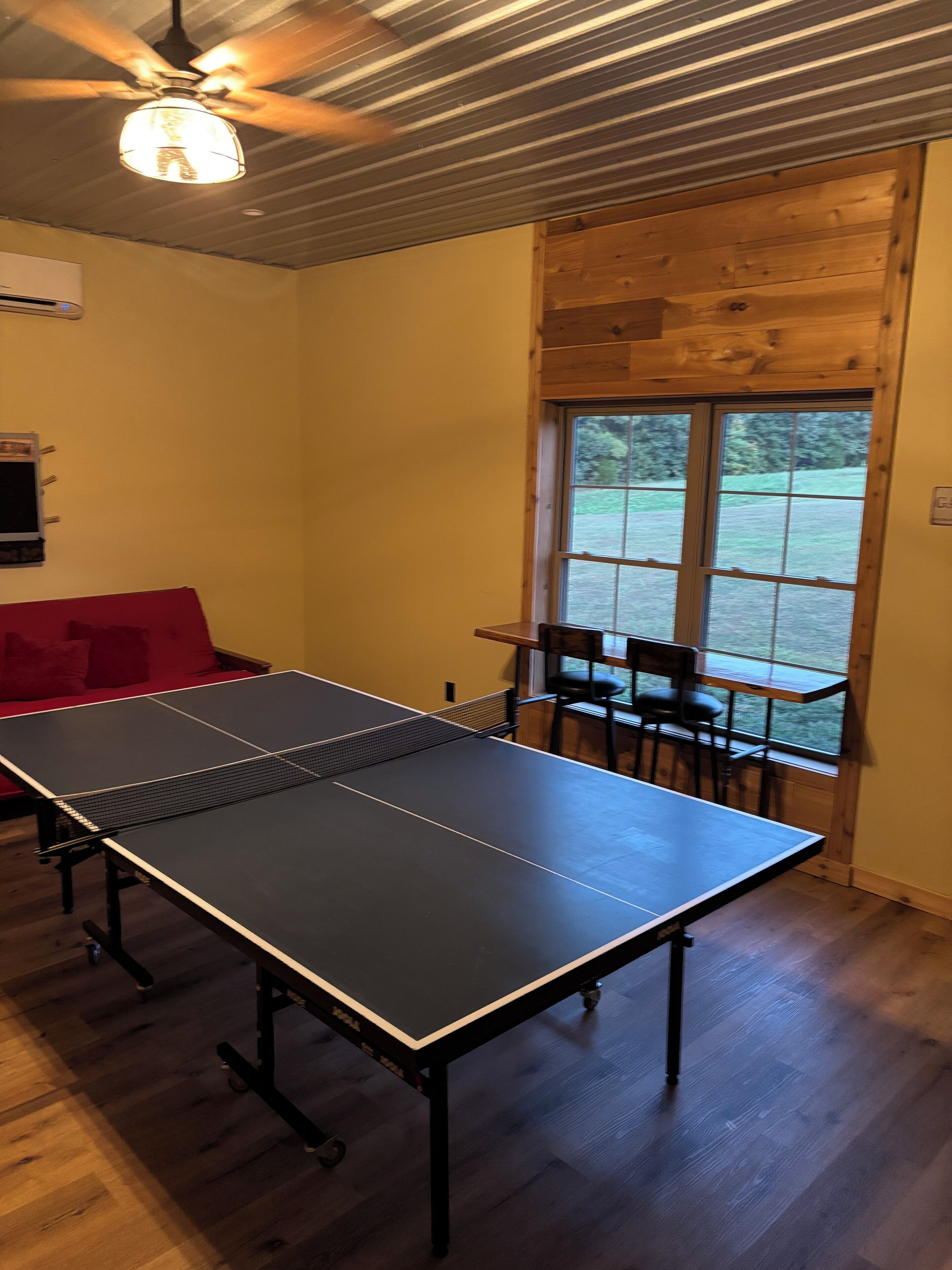 A ping pong table in a room with wooden flooring, a ceiling fan, and a window with a view of greenery.