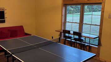 A ping pong table in a room with wooden flooring, a ceiling fan, and a window with a view of greenery.