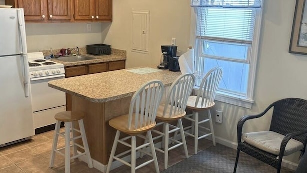 A kitchen with wooden cabinets, a white refrigerator, a stove, and a bar area with four white chairs.