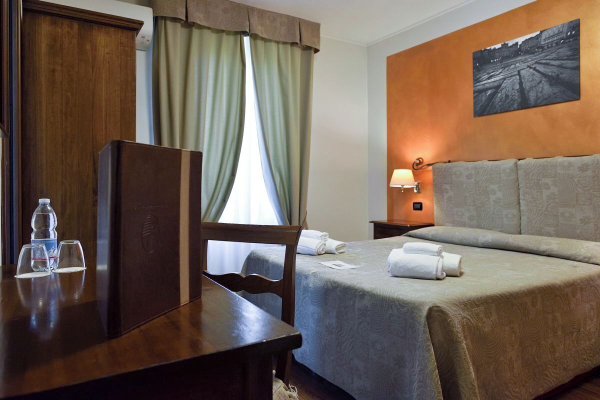A hotel room with a bed, a desk with a water bottle and glasses, a lamp, and a cityscape painting on the wall.