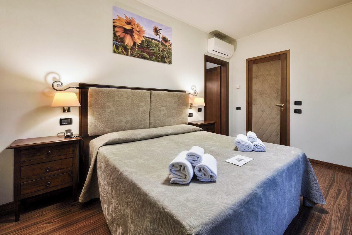 A hotel room with a bed, two bedside tables, a wall-mounted air conditioner, a door, and a painting of flowers.
