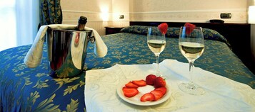 Un letto con una coperta blu a motivi floreali, due bicchieri di vino bianco e un piatto di fragole su un tavolo.