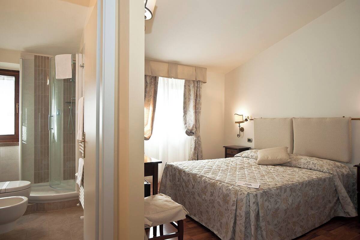 Un dormitorio con cama, mesita de noche, cortinas en la ventana y zona de ducha con puerta de cristal.
