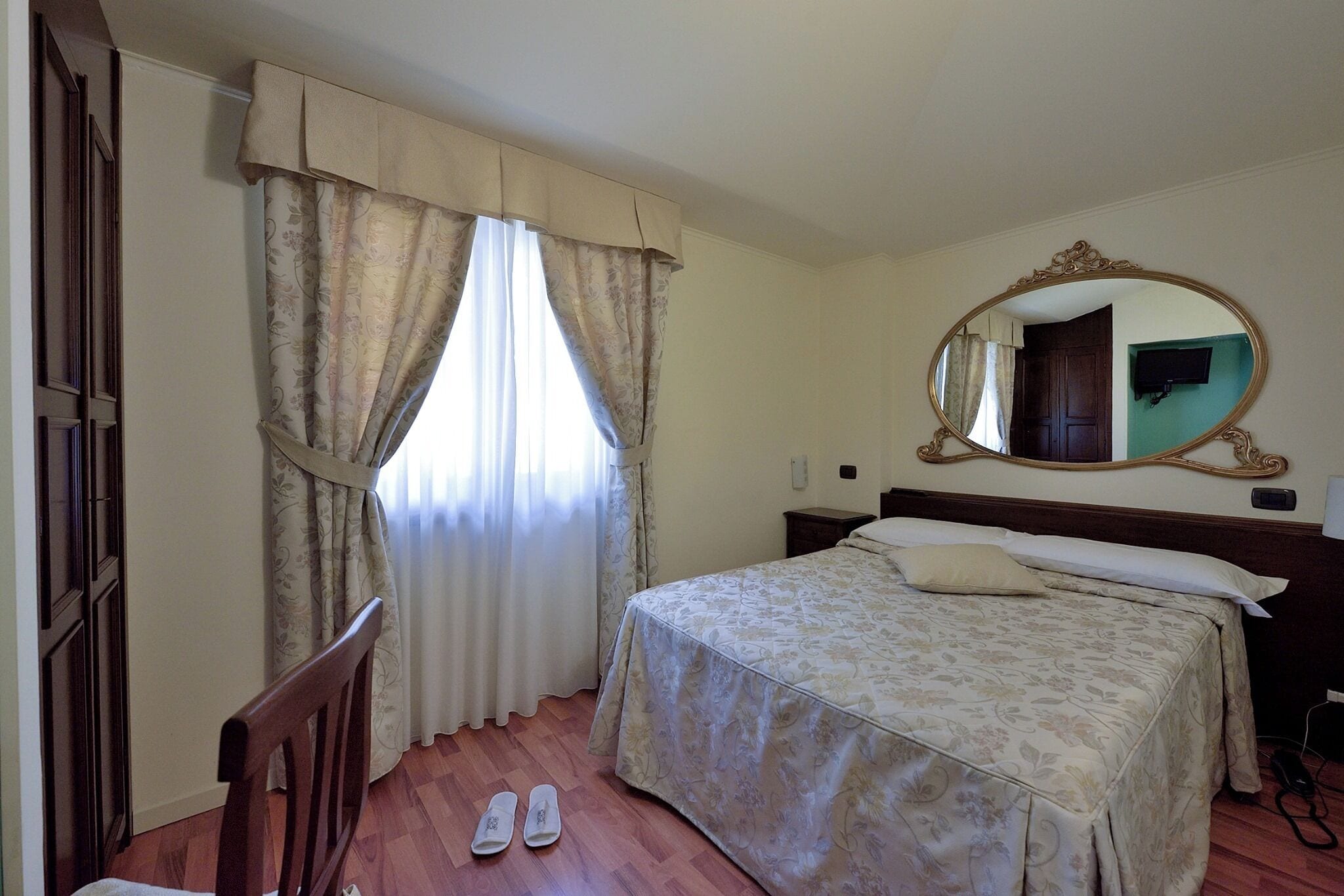 A bedroom with a bed, a mirror, a chair, and a pair of slippers on the floor.