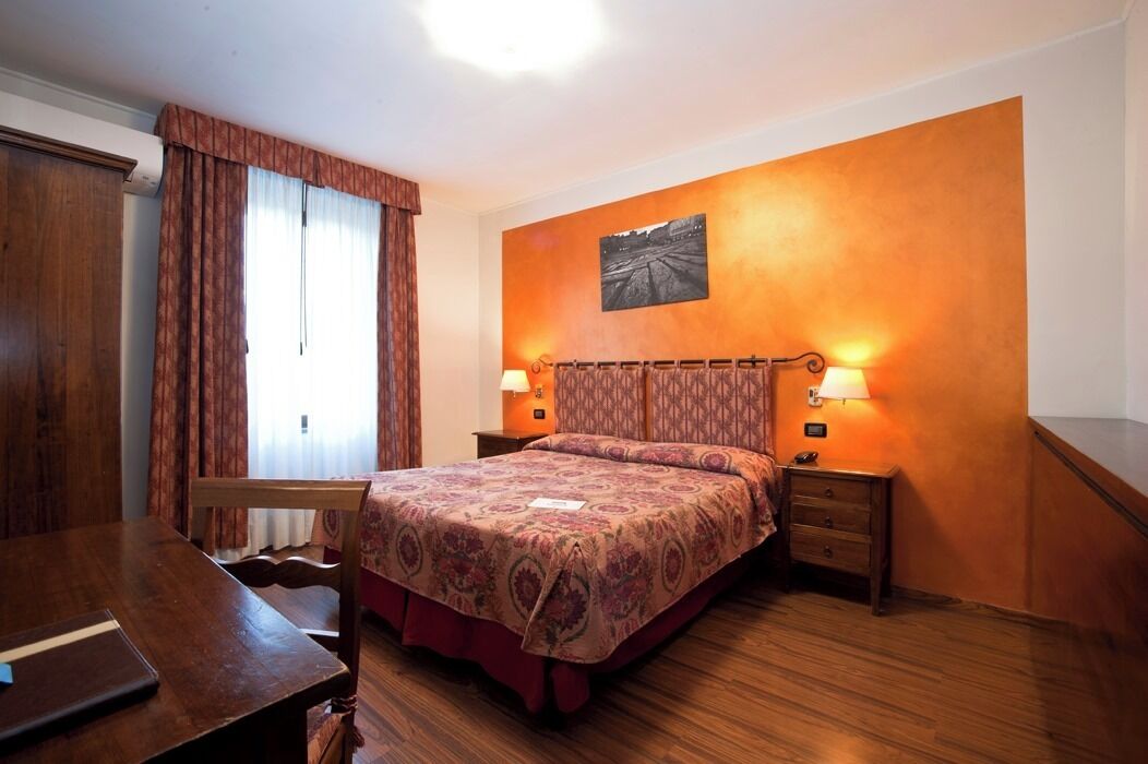 A bedroom with a wooden bed, a desk, and a chair. There are two bedside lamps and a wall-mounted picture.