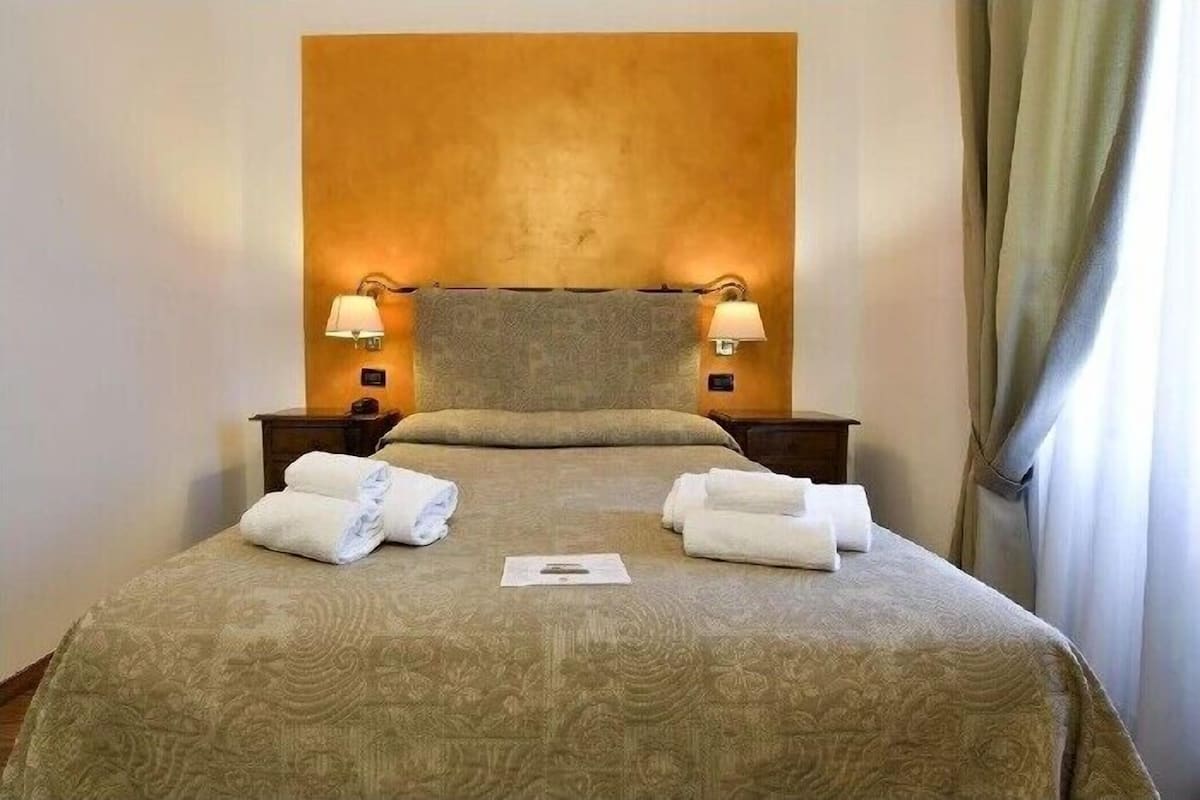 Una cama bien hecha con una colcha estampada, dos lámparas de noche y toallas dobladas sobre la cama.