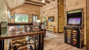 Ein gemütliches Blockhäuschen mit Holzwänden und Deckenventilator. Eine Kaffeestation mit einer Keurig-Maschine und einem Schild mit der Aufschrift "Coffee Bar" ist zu sehen. Außerdem steht ein Arcade-Automat mit der Bezeichnung "Legends Ultimate" dort.