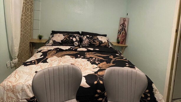 Un dormitorio con cama, dos sillas, ventilador de techo, mesita de noche y un cuadro en la pared.