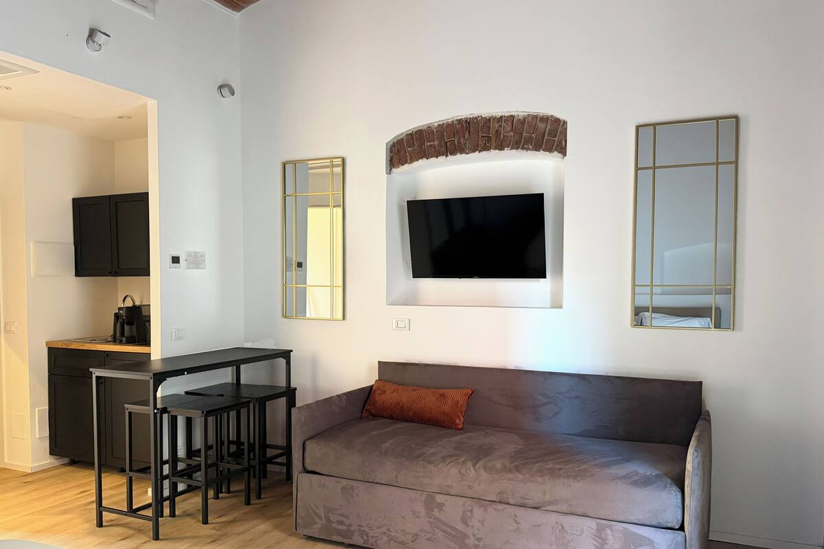 Una sala de estar moderna con un sofá gris, un televisor montado encima y una pequeña zona de cocina con una cafetera.