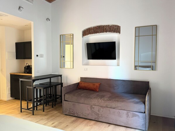 A modern living room with a grey sofa, a TV mounted above it, and a small kitchen area with a coffee machine. - Palazzo Garbusi (Sarzana)