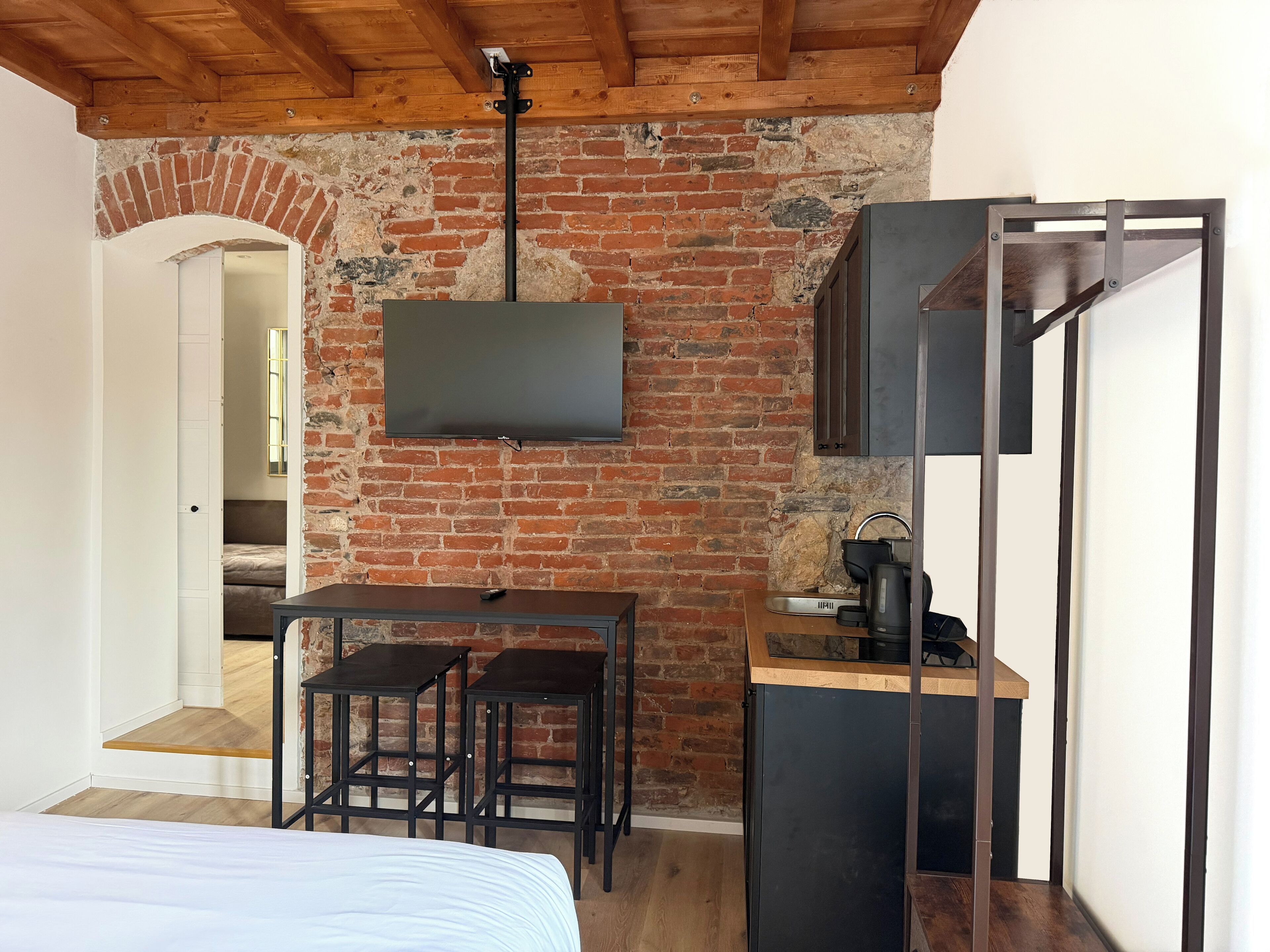 Een knus, compacte kamer met een bakstenen muur, een flatscreen televisie boven een houten tafel met twee krukken, een bed met een wit hoofdeinde, en een kleine kitchenette met een koffiezetapparaat.