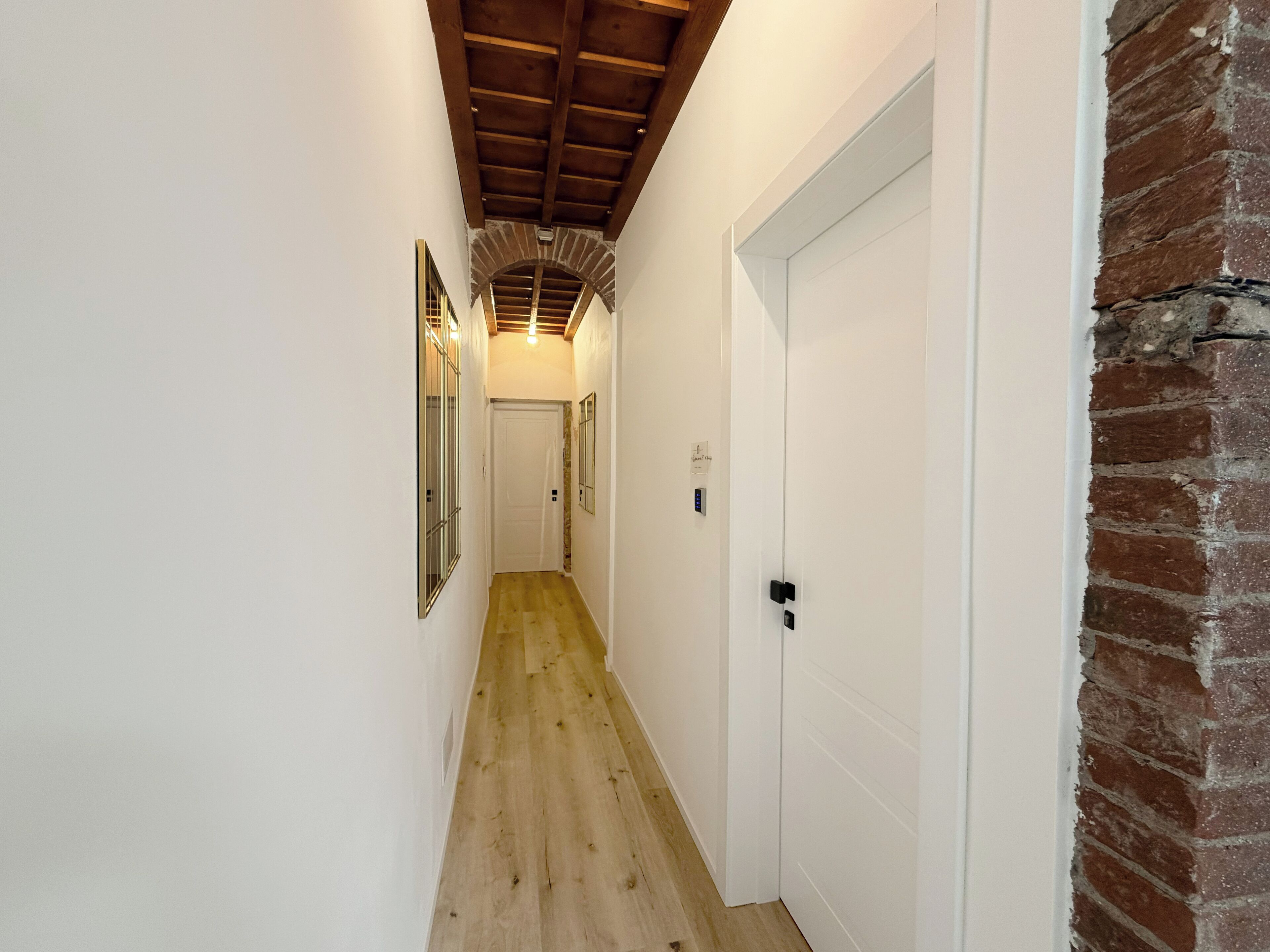 Un pasillo estrecho con suelo de madera, paredes blancas y una pared de ladrillo a la derecha. Hay dos puertas y un pequeño ventanal con marco metálico.