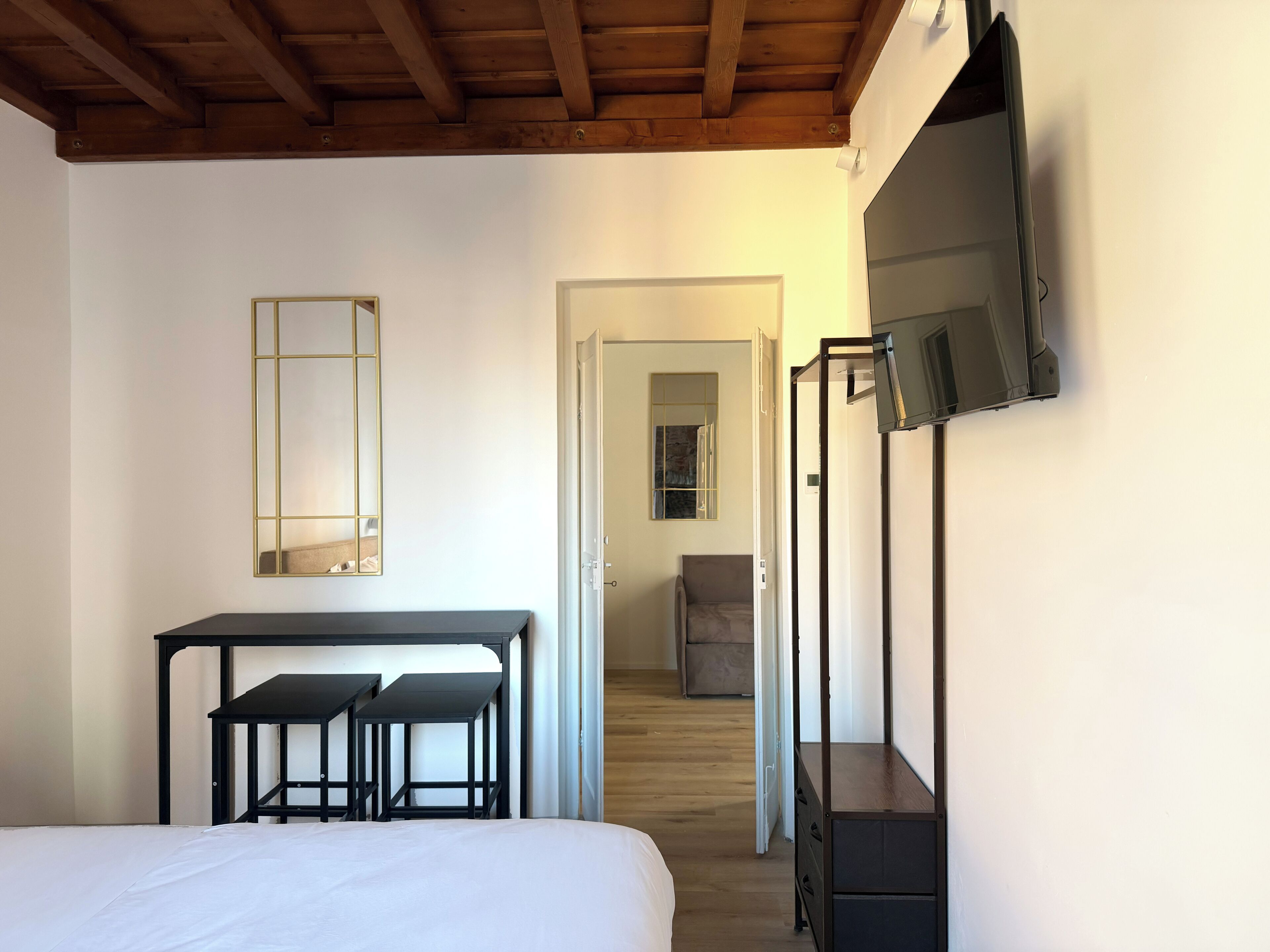 Un dormitorio con cama, escritorio y dos sillas, televisor montado en la pared, y puerta que da a otra habitación.