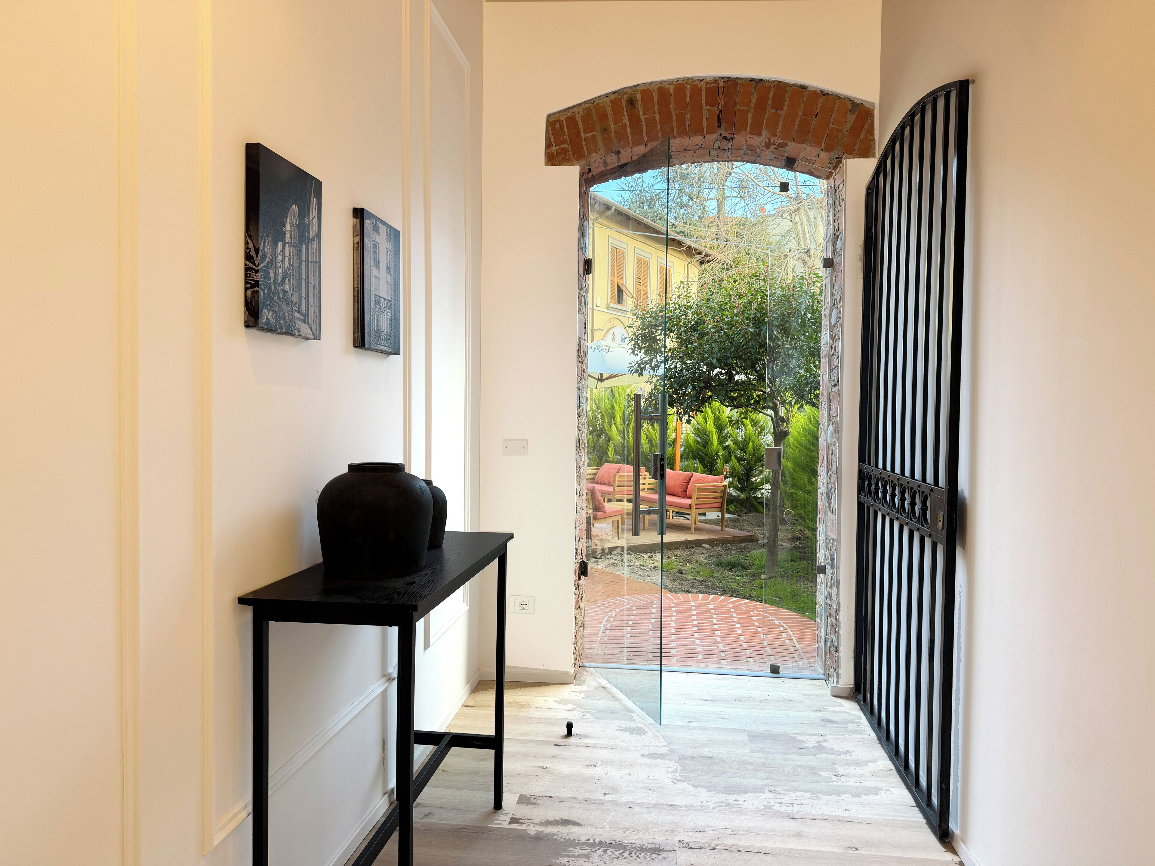 A hallway with a black table, two framed pictures, and a glass door leading to an outdoor seating area with a red bench and greenery.