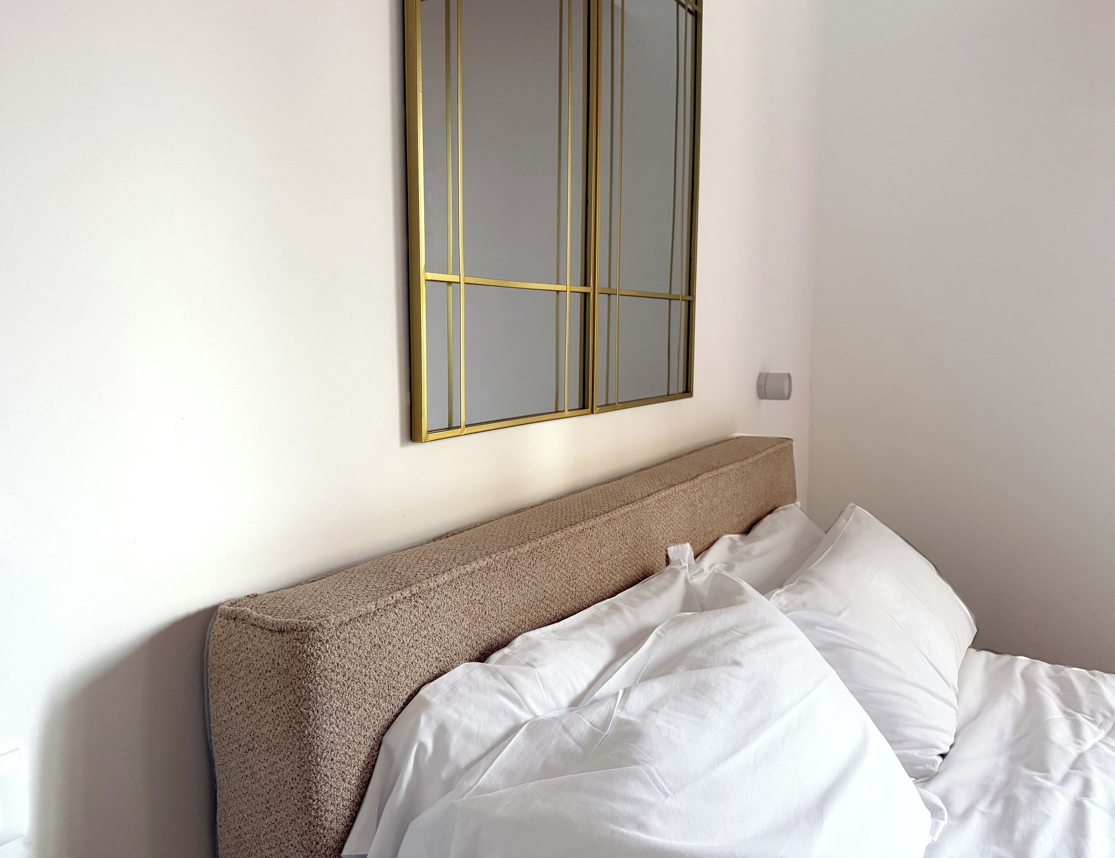 A neatly made bed with white linens and a brown headboard, a wall-mounted mirror with a gold frame, and a small wall-mounted light fixture.