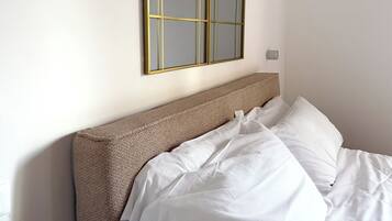 A neatly made bed with white linens and a brown headboard, a wall-mounted mirror with a gold frame, and a small wall-mounted light fixture.