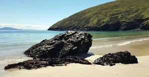 A beach with large rocks, clear water, and a green hill in the background.