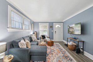 Ein Wohnzimmer mit grauem Sofa, einem Couchtisch und einem gemusterten Teppich. An der Wand hängt ein Fernseher und im Hintergrund ist eine Tür zu sehen.