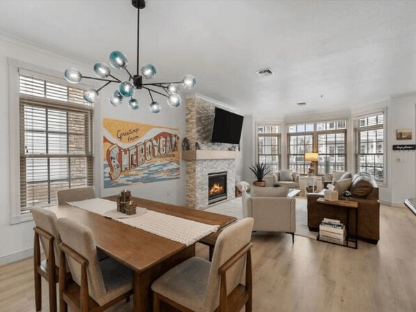 A spacious living room with a fireplace, a dining area with a wooden table and chairs, and large windows with blinds.