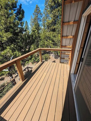 Une terrasse en bois, dotée de rampes de protection et entourée d’arbres, mène à un bâtiment avec une porte en verre.