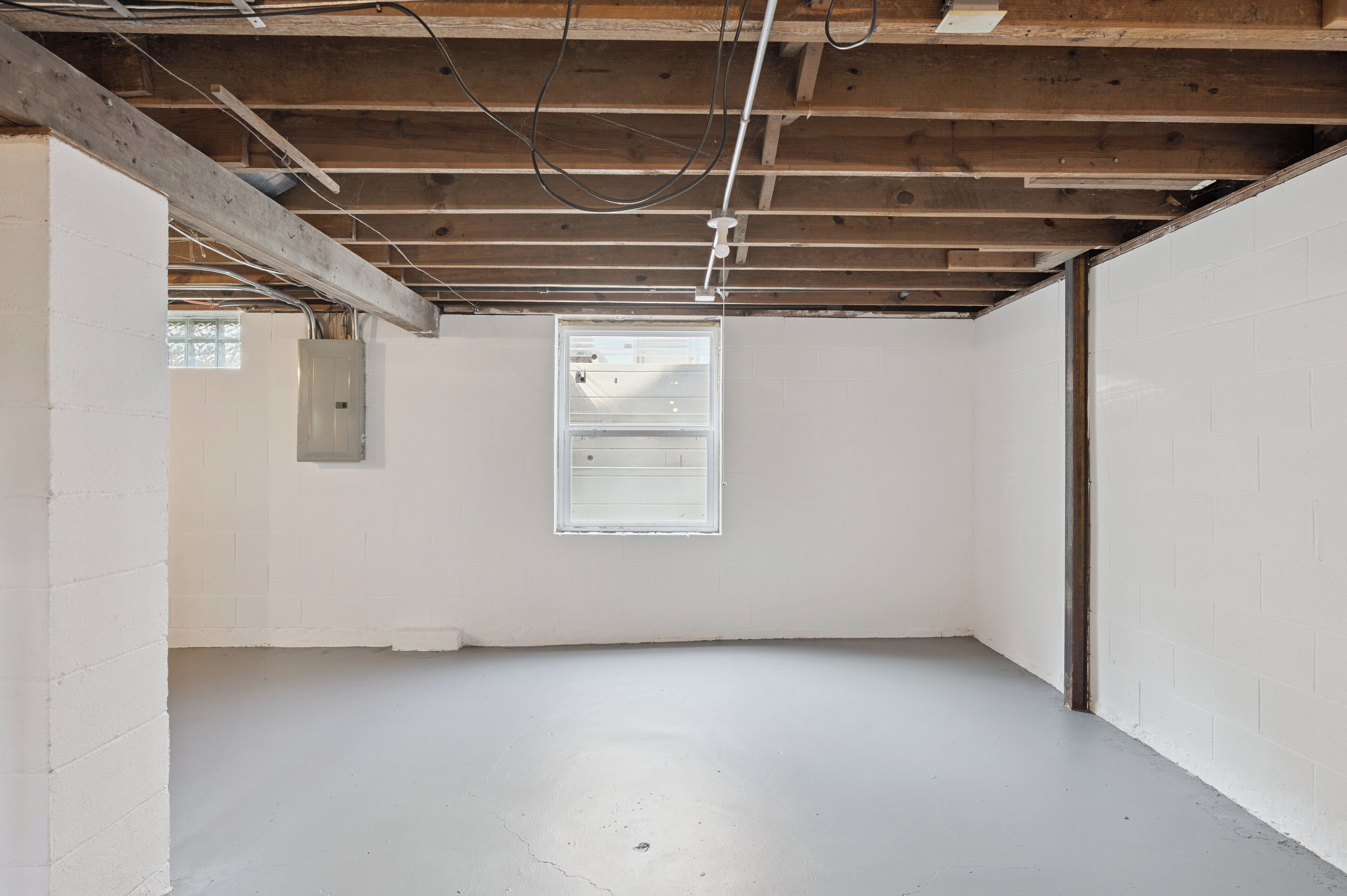 Una habitación con paredes blancas, piso de concreto y vigas de madera a la vista en el techo.