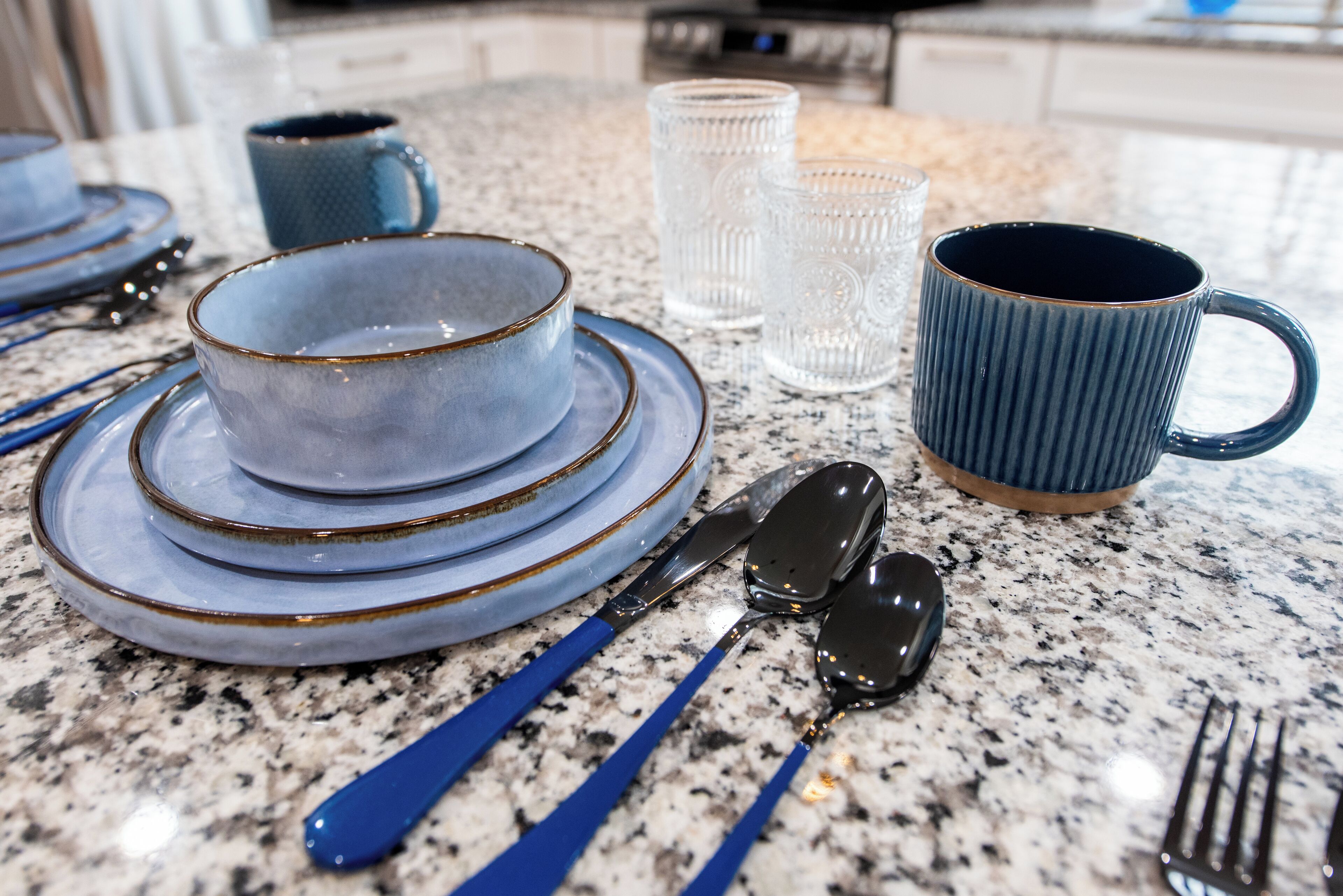 A kitchen counter with stacked plates, a blue mug, clear glasses, and cutlery.