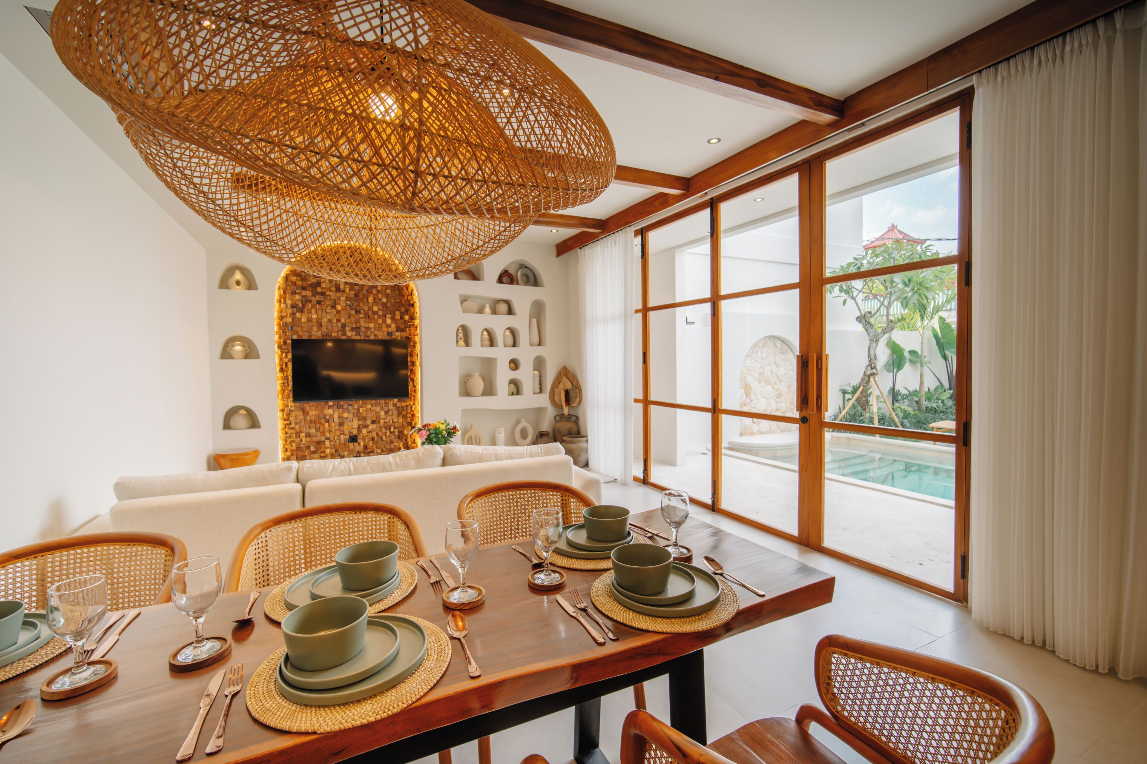 Comedor con una mesa de madera dispuesta para cuatro, una lámpara de techo tejida y vista a una piscina a través de amplios ventanales.