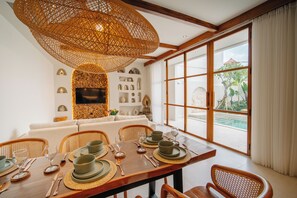 A dining area with a wooden table set for four, a woven ceiling light, and a view of a swimming pool through large windows. - Root Bumbak Bali (Kerobokan)
