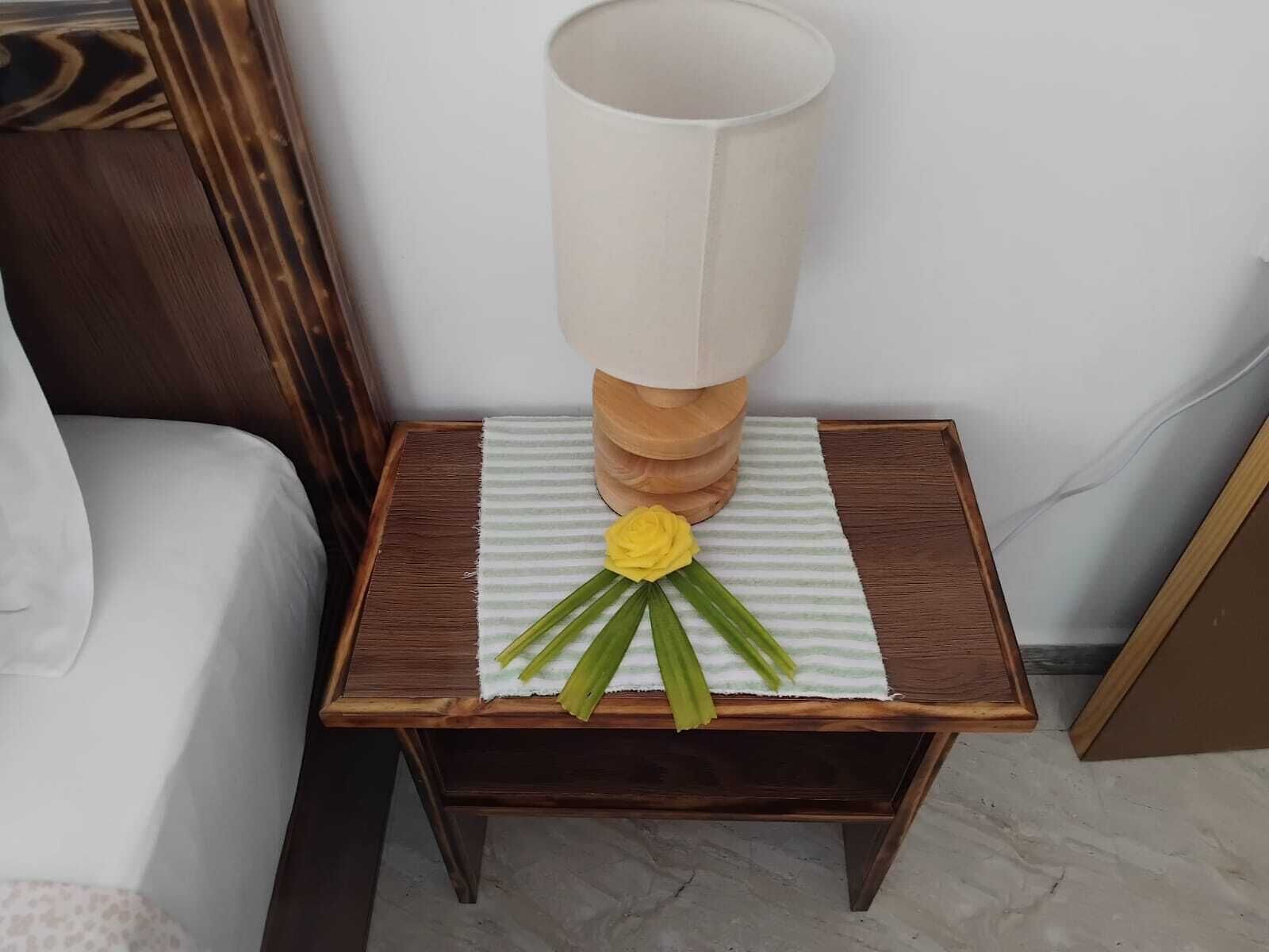 Una mesita de noche de madera con una lámpara y una flor amarilla sobre un tejido a rayas.