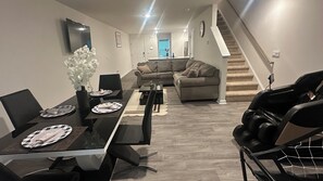 A modern living room with a dining area, a grey sofa, a staircase, and a flat-screen TV mounted on the wall.