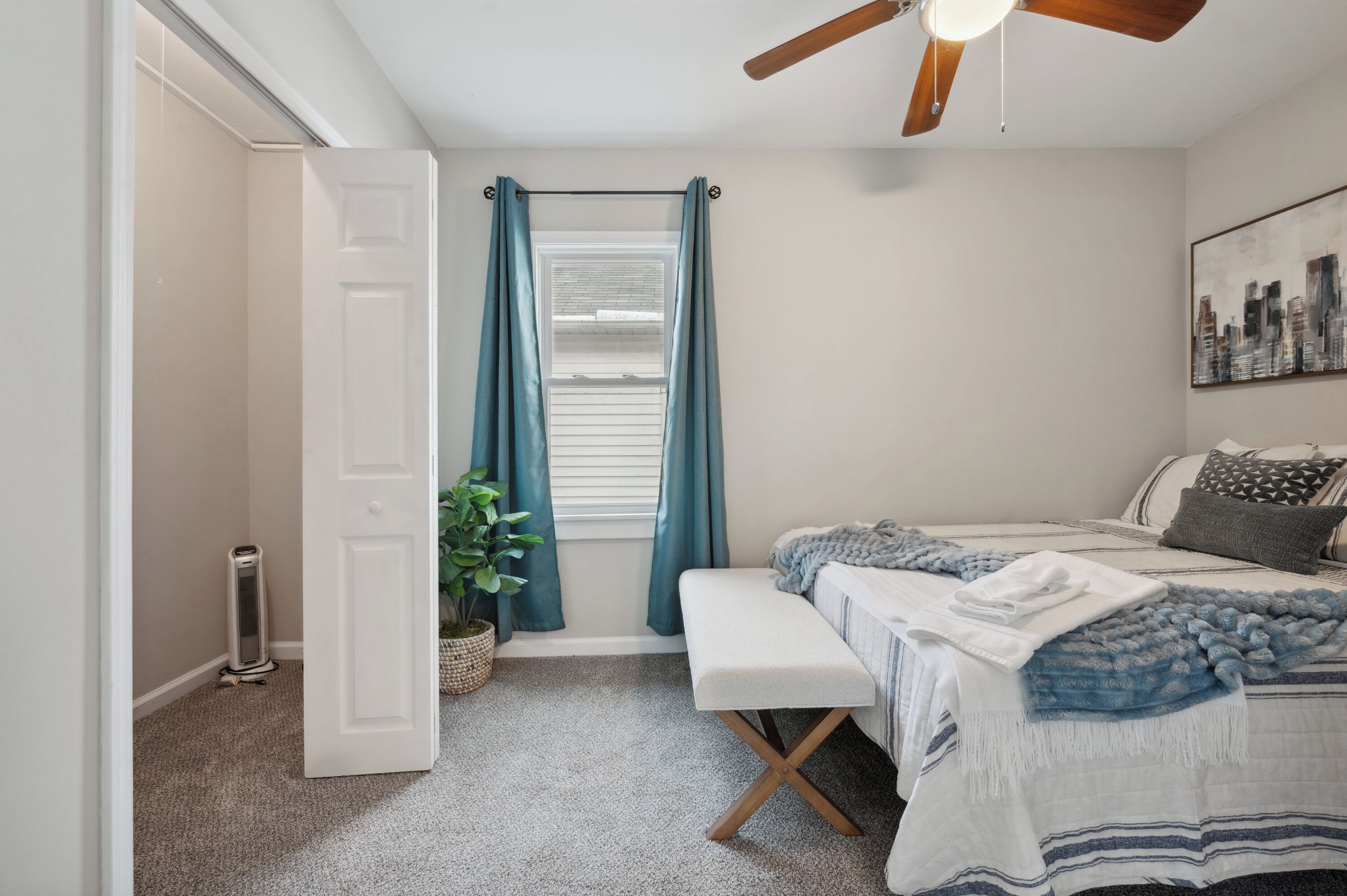 A bedroom with a bed, a bench, a ceiling fan, a window with curtains, and a cityscape painting on the wall.