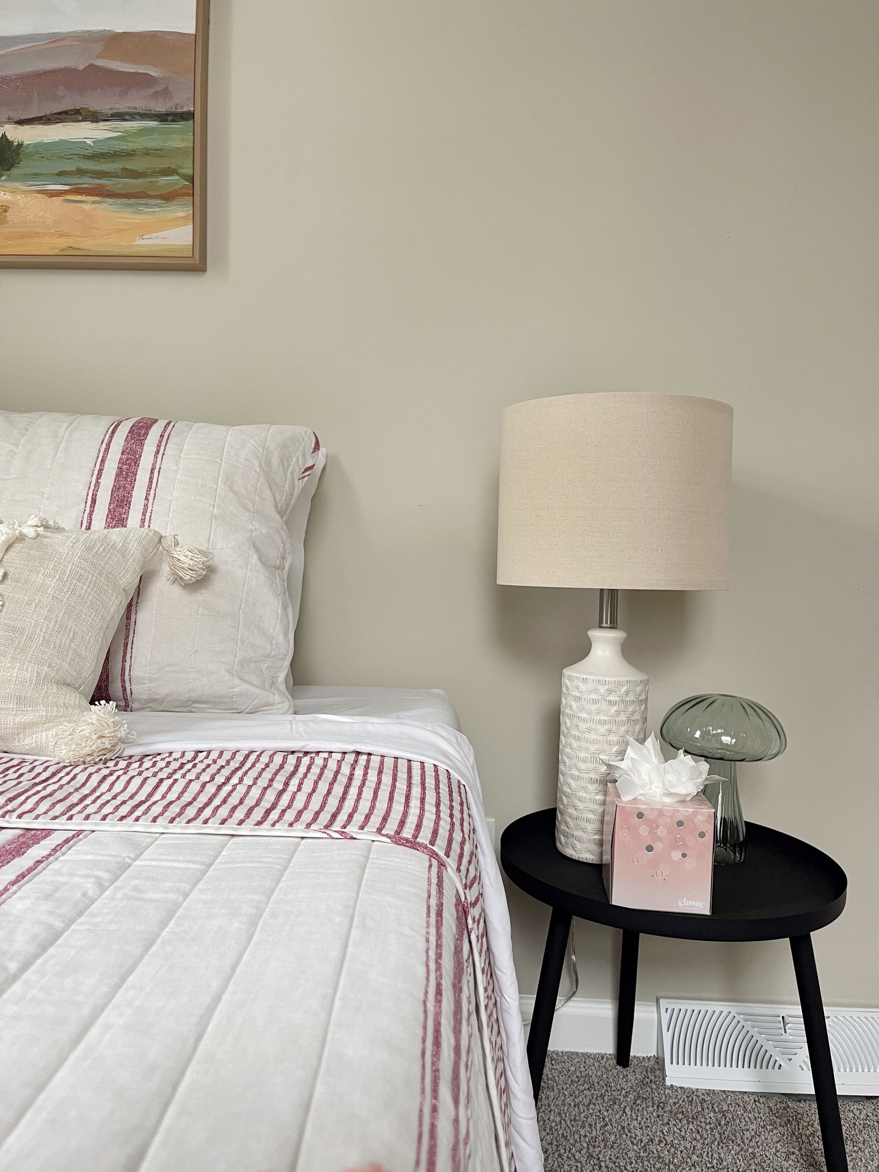A neatly made bed with a striped bedspread, a white pillow with tassels, and a bedside table with a lamp, a decorative box, and a vase.