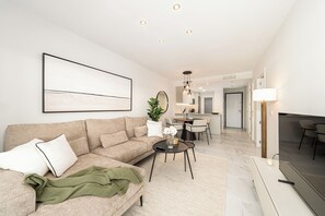 Ein modernes Wohnzimmer mit einer Couch, einem Couchtisch und einem Fernseher. Im Hintergrund befindet sich ein Essbereich mit Stühlen und einem Tisch.