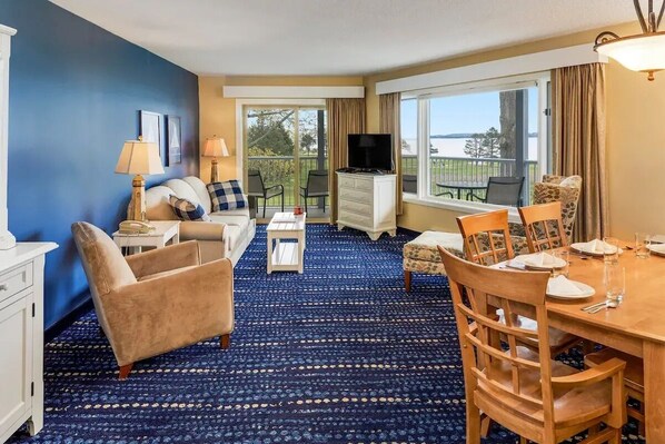 A hotel room with a blue carpet, a sofa, a TV, a dining table set for four, and a balcony with a view of the outdoors.