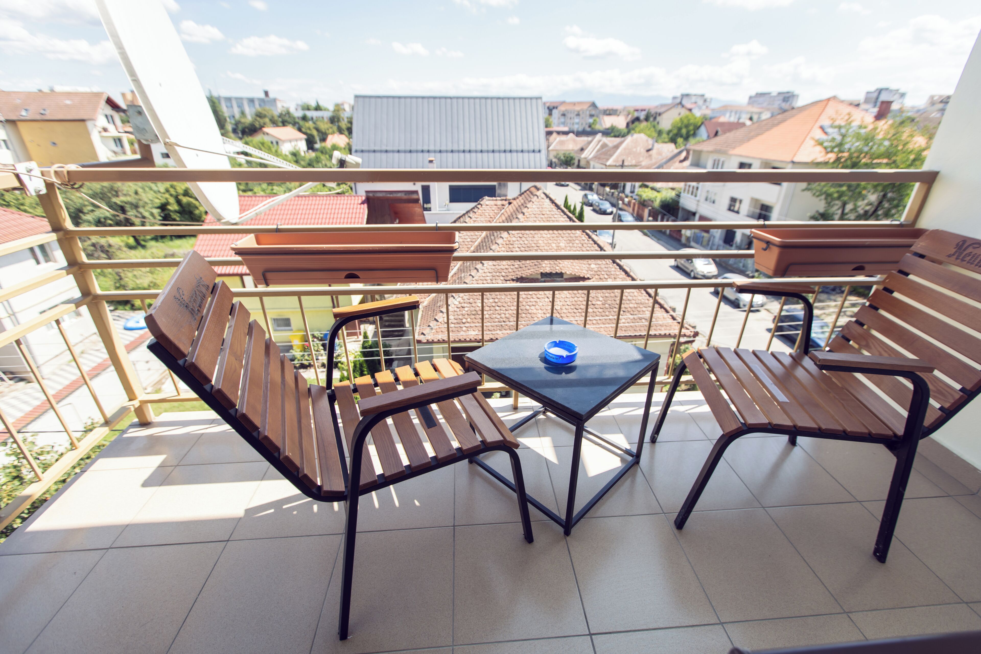 A balcony with two wooden chairs and a small table, overlooking a residential area with houses and a clear sky.