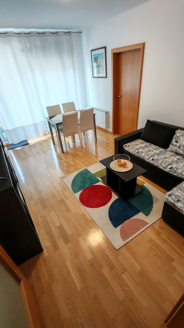 A living room with a sofa, a coffee table, a dining table with chairs, a door, and a radiator.